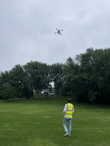 Drone pilot wearing a safety vest controls a commercial drone flying overhead during an aerial survey